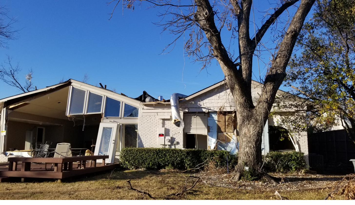 Leo's family home in Dallas heavily damaged by a tornado, with the roof torn away and debris scattered under a clear blue sky